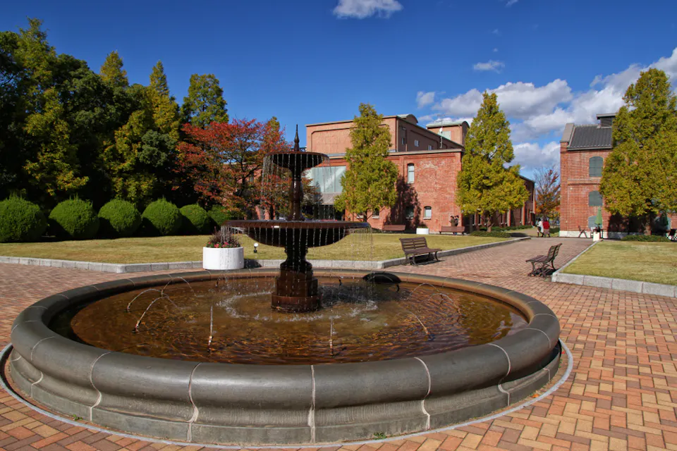 一个圆形喷泉，水流从多个层次倾泻而下，位于砖铺院子的中心，周围是绿色的树木和灌木。在背景中是红砖建筑，天空晴朗，飘着几朵散云。