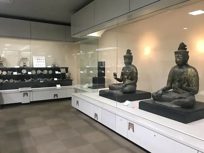 一个博物馆展览展示了两个大型坐姿佛像，它们被玻璃罩住，放置在白色平台上。在背景中，有架子展示各种文物，包括陶瓷盘和陶器。房间灯光明亮，设计现代。