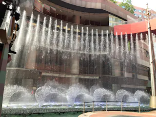 A large fountain shoots arcs of water upward in front of a modern, curved building with glass and red accents. Sunlight highlights the spray, and some greenery is visible above the structure.