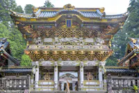 Elaborate, multi-tiered wooden shrine structure adorned with intricate carvings and gold accents, showcasing traditional Japanese architectural elements. Stone steps lead to the entrance, guarded by detailed animal and deity sculptures, set against a backdrop of dense green trees.