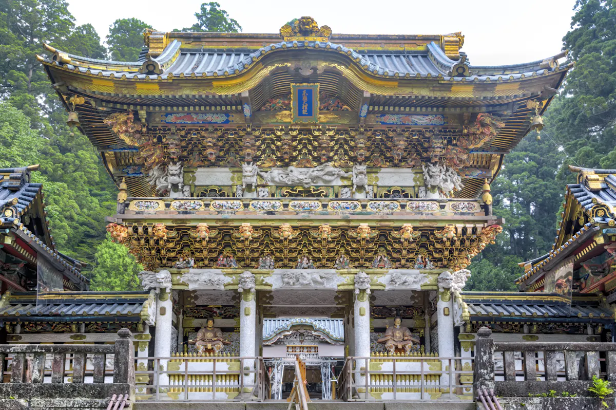 Elaborate, multi-tiered wooden shrine structure adorned with intricate carvings and gold accents, showcasing traditional Japanese architectural elements. Stone steps lead to the entrance, guarded by detailed animal and deity sculptures, set against a backdrop of dense green trees.