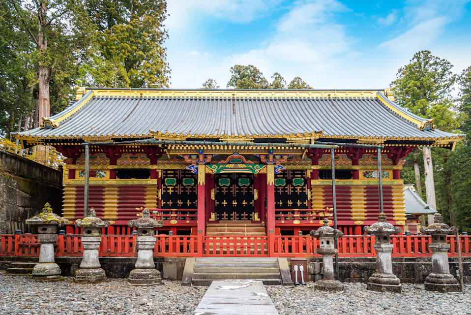 色とりどりで装飾豊かな神社が、明るい青空の下で高くそびえる大きな木々に囲まれています。神社には、赤、金、緑の華麗な彫刻や装飾が施され、入口まで続く道沿いには伝統的な石灯篭が並んでいます。