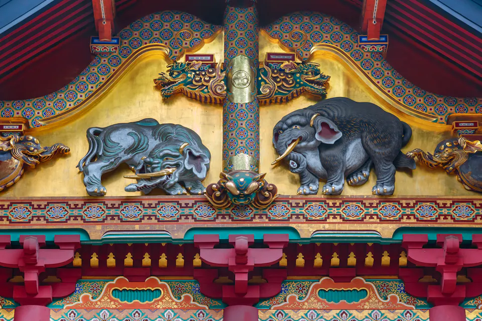 伝統的でカラフルなアジアの建物のファサードを飾る2頭の象の詳細な木彫り。彼らは鮮やかな色と緻密なパターンで彩色されており、赤、青、金、緑の装飾的なデザインやモチーフに囲まれています。