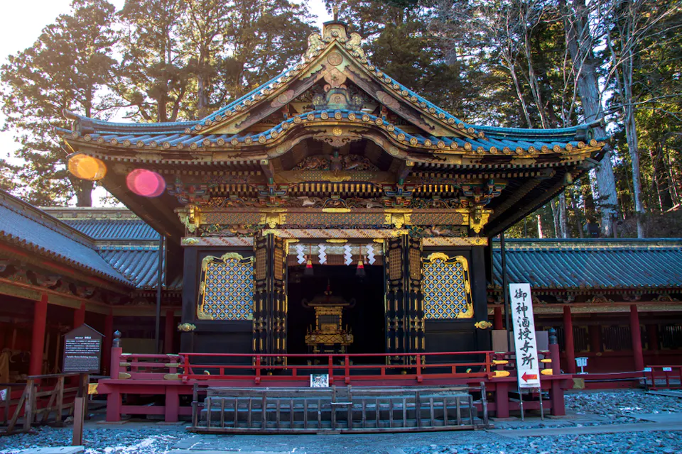 金色と黒のファサードを持つ装飾的な日本の寺院の建物が高い木々に囲まれて立っている。建築の詳細には精巧な彫刻と鮮やかな色が含まれている。右側には日本語の文字がある看板がある。地面は砂利と石で覆われている。