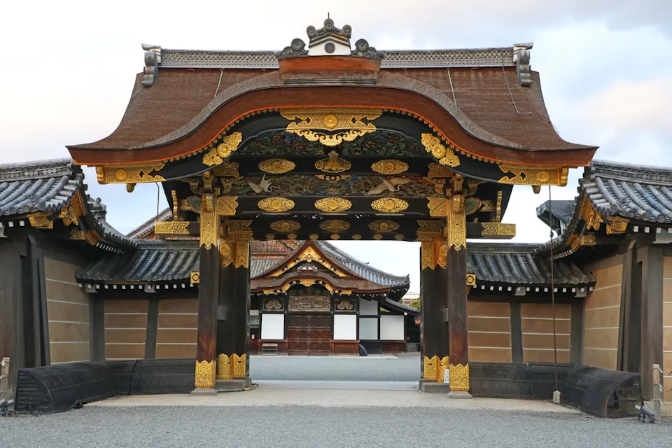 複雑な建築詳細を持つ伝統的な日本の寺院が、部分的に曇った空の背景に立っています。寺院には、追加の寺院構造に囲まれた中庭に通じる目立つアーチウェイがあります。