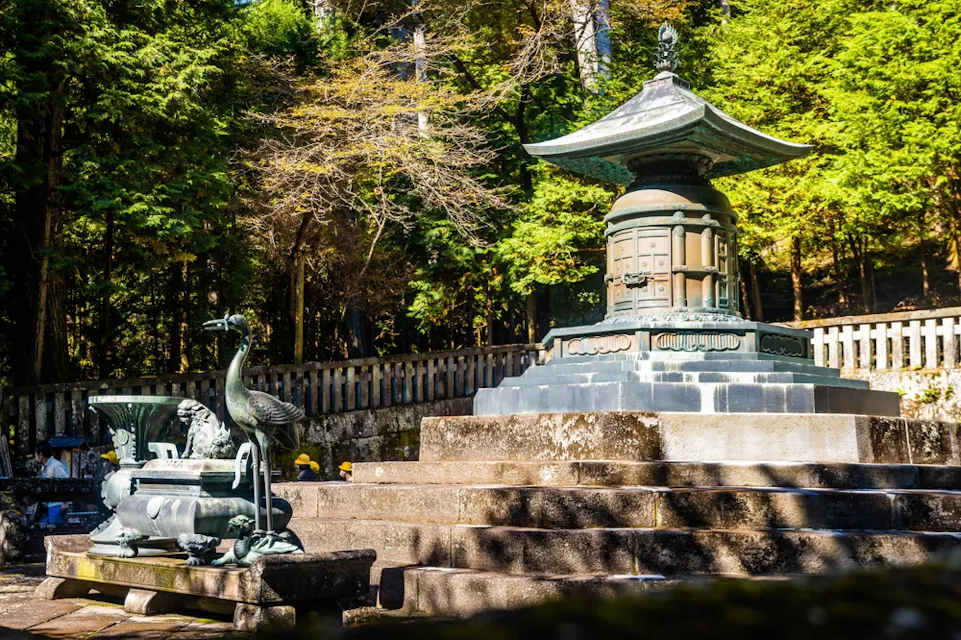 荘厳なモニュメントが石のプラットフォームの上に立っており、緑豊かな木々を背景にしている。左側には、装飾的な台座の上に飾られた鳥の彫像が置かれ、シーンの静かで歴史的な雰囲気を増している。