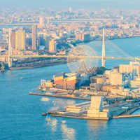 Osaka Bay Area Aerial view of a coastal cityscape featuring a large Ferris wheel near a suspension bridge. Numerous buildings are clustered around the waterfront. The sunlight casts a warm glow over the city, highlighting the calm waters of the surrounding bay.