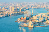 Aerial view of a coastal cityscape featuring a large Ferris wheel near a suspension bridge. Numerous buildings are clustered around the waterfront. The sunlight casts a warm glow over the city, highlighting the calm waters of the surrounding bay.