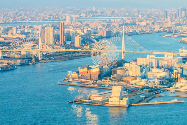 Aerial view of a coastal cityscape featuring a large Ferris wheel near a suspension bridge. Numerous buildings are clustered around the waterfront. The sunlight casts a warm glow over the city, highlighting the calm waters of the surrounding bay.