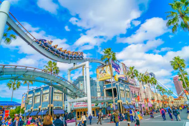 People walk beneath a roller coaster at an amusement park. The coaster has a white track and a blue train filled with riders. The park features colorful buildings and palm trees under a partly cloudy blue sky. The scene is lively and vibrant.