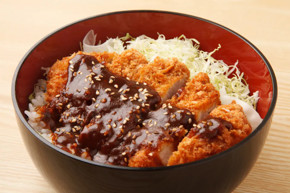 Miso Katsu A bowl of Japanese katsu-don, featuring breaded and fried pork cutlet drizzled with a thick, dark sauce, sprinkled with sesame seeds, on a bed of shredded cabbage and rice. The dish is served in a black bowl with a red interior, placed on a light wood surface.