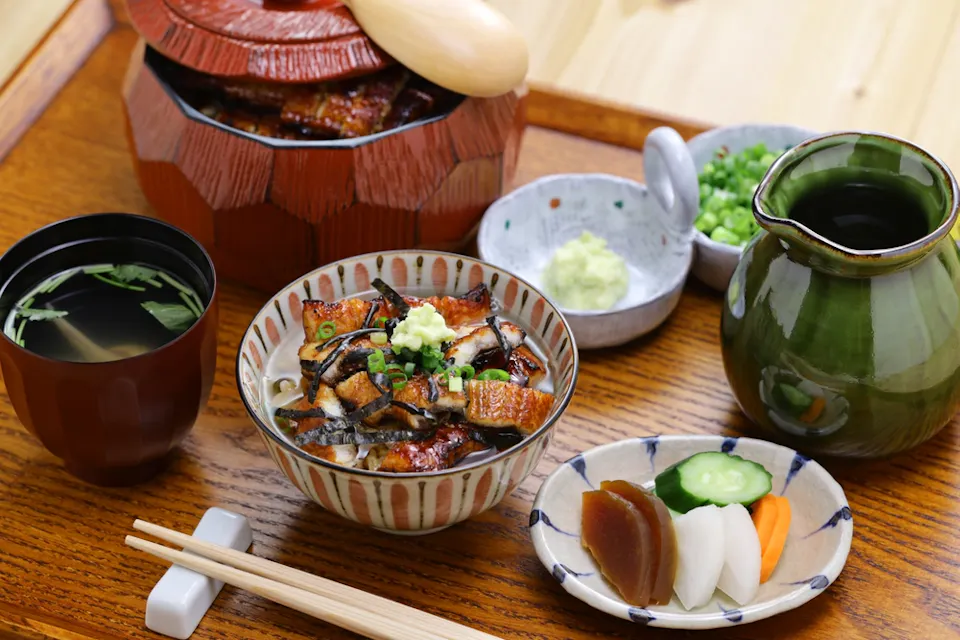 A wooden tray holds a Japanese meal consisting of a bowl of rice topped with grilled eel, seaweed, and green onions. Surrounding it are a bowl of soup, pickled vegetables, a small dish of wasabi, a pot of green tea, green onions, and a wooden spoon.