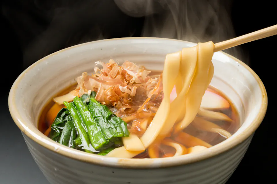 Kishimen A steaming bowl of udon noodle soup with thick noodles being lifted by chopsticks. The soup features vibrant green leafy vegetables, umami-rich broth, and bonito flakes. The dish is served in a ceramic bowl against a dark background.