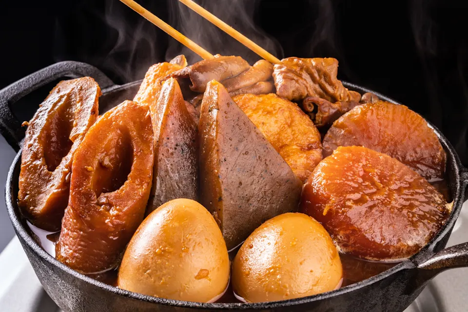 A hot pot filled with an assortment of simmered Japanese oden ingredients, including fish cakes, boiled eggs, daikon radish, konjac, and other delicacies, all in a savory broth. Chopsticks are resting on top, with steam rising from the meal.