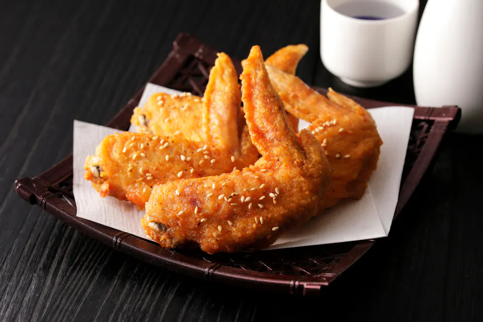 A plate of golden, crispy fried chicken wings topped with white sesame seeds is served on a paper-lined tray. In the background, there are two small white cups, possibly for sake or tea, on a dark wooden surface.