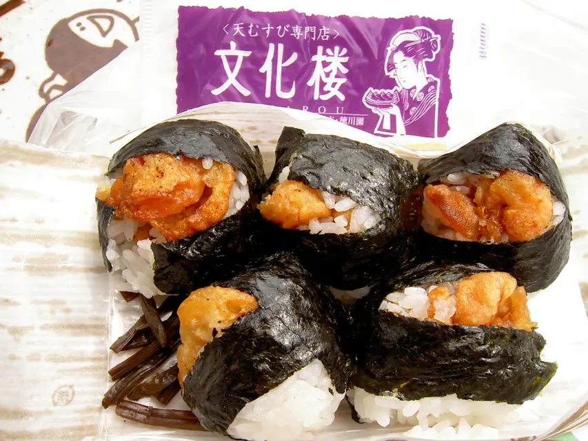 A plastic container holds five onigiri, each wrapped in seaweed and filled with crispy fried chicken. Behind the onigiri, there is a partially visible white and purple package that features an illustration of a traditional Japanese woman.