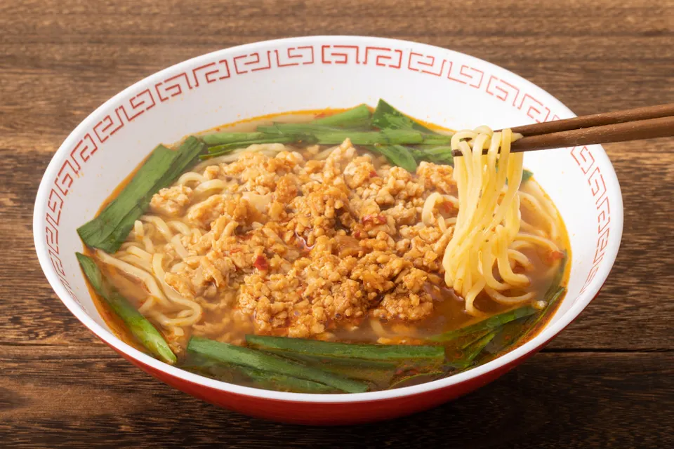Taiwan Ramen A bowl of ramen with a rich broth, topped with ground meat and green vegetables, placed on a wooden table. A pair of chopsticks holds a portion of noodles above the bowl. The bowl has a traditional red and white patterned rim.
