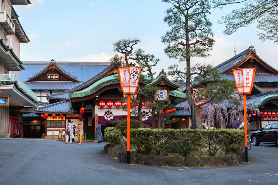 Gion Corner 一座传统的日本建筑,华丽的屋顶结构被红色灯笼照亮,伫立在修剪整齐的花园和铺成的车道后面。前景中有两个带有日文的橙色指示牌,两个身影在入口附近可见。