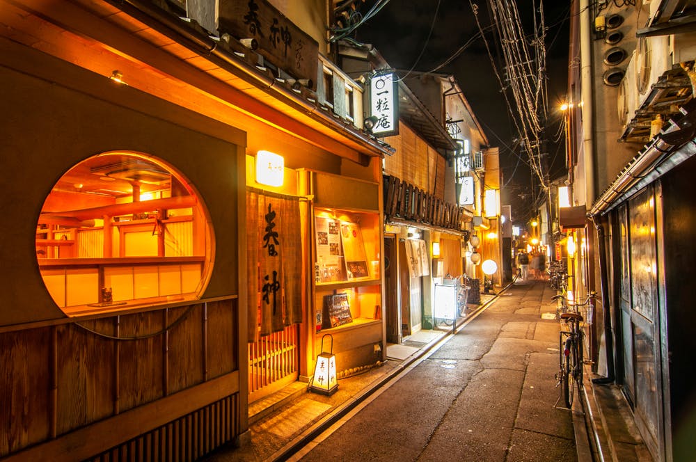 A quiet, narrow street in a Japanese town at night, illuminated by warm lights from cozy wooden shops and restaurants. A round window is prominently featured on one building. Overhead, utility wires crisscross, and bicycles are parked along the sidewalk.