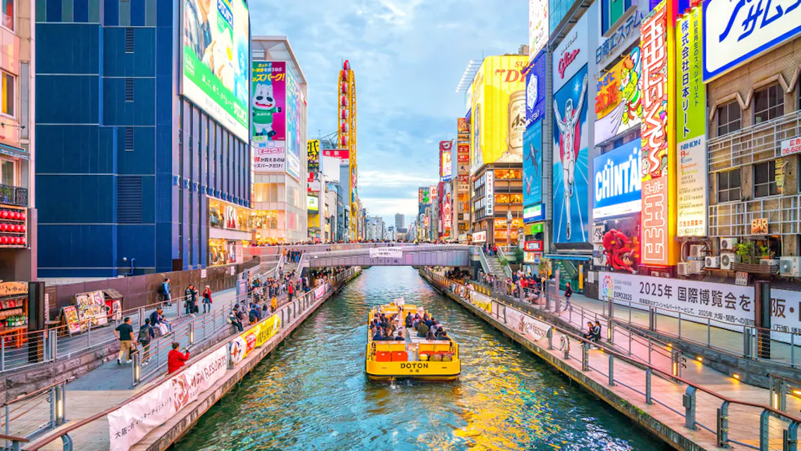 Dotonbori 一幅生动的城市水道场景,一艘黄色船只载着乘客在运河上行驶。高楼大厦装饰着五彩缤纷的广告和广告牌,沿着运河两侧排成一排。行人沿着桥和河岸散步,享受着热闹的氛围。