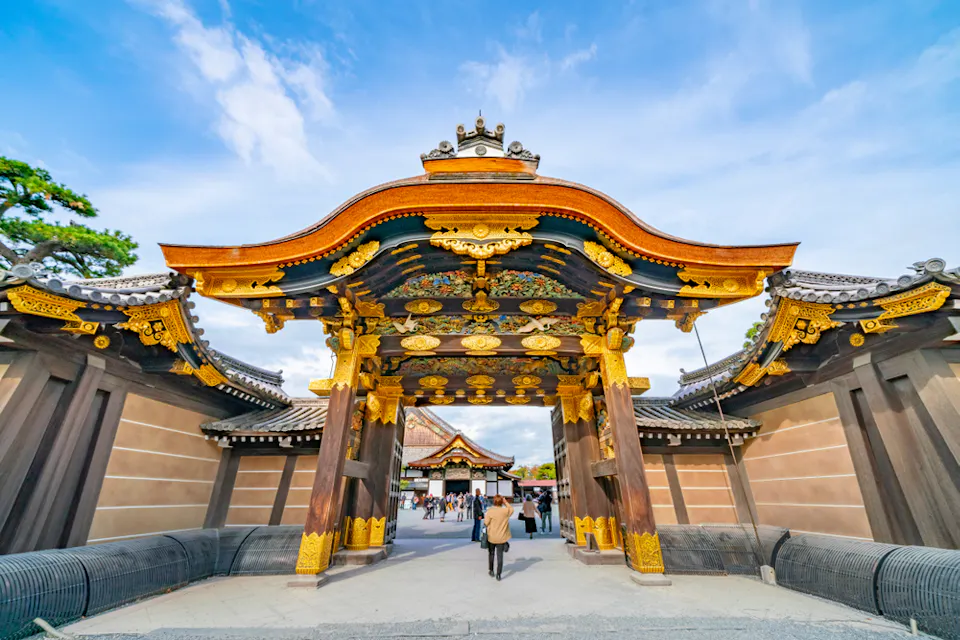 Kyoto Imperial Palace or Kyoto Gosho 一个人站在装饰有复杂金色细节和华丽雕刻的传统日本木制大门前。大门通向一个庭院,背景中有其他游客,在明亮的蓝天和散布的白云下。