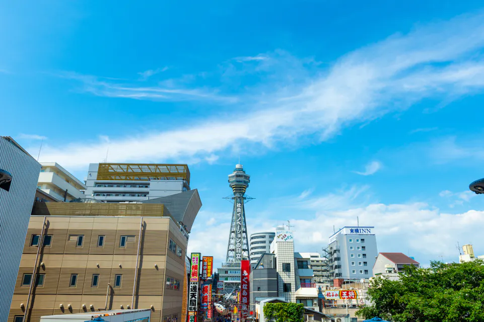 Tsutenkaku Tower 在一个晴朗的日子里,展示大阪通天阁的城市景观。天际线包括各种建筑、带有日文的标志,以及前景中的几棵绿树。天空是明亮的蓝色,飘着零星的云朵。