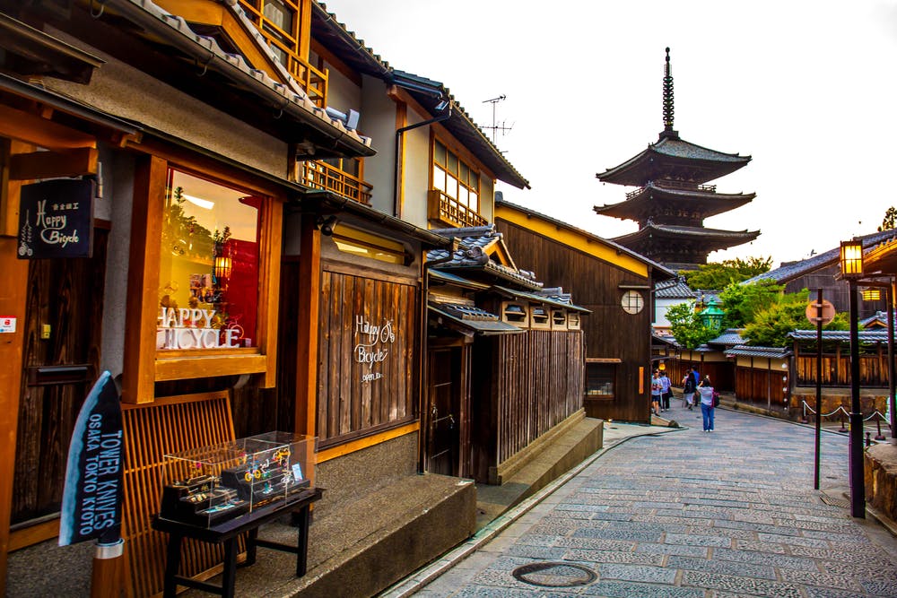 A charming street scene in Kyoto, featuring traditional wooden buildings with tiled roofs. A shop displays a "Happy Bicycle" sign. In the background, a tall pagoda rises above the street while a few pedestrians stroll along the cobblestone path, enjoying the serene atmosphere.