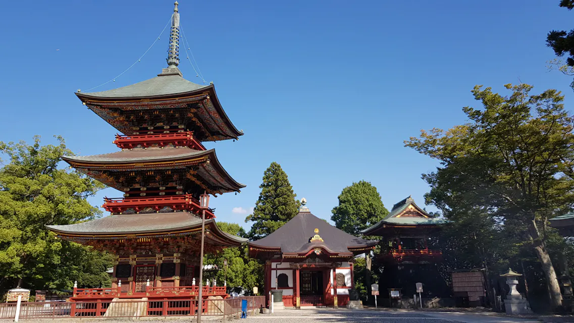 A traditional Japanese temple complex set against a clear blue sky. It features a multi-tiered pagoda to the left, a main hall in the center with a curved roof, and another structure to the right. The area is surrounded by lush green trees.
