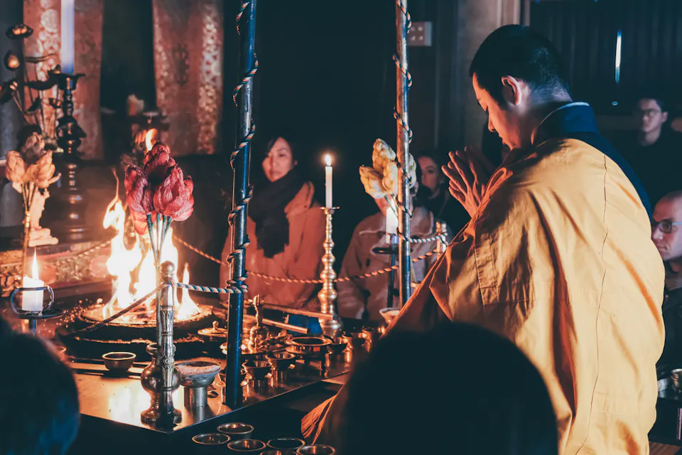 A monk, dressed in yellow robes, performs a ritual in a dimly lit temple. He's surrounded by various items, including a candle, flowers, and metal tools, while several people sit in the background, observing the ceremony. The atmosphere is solemn and reverent.