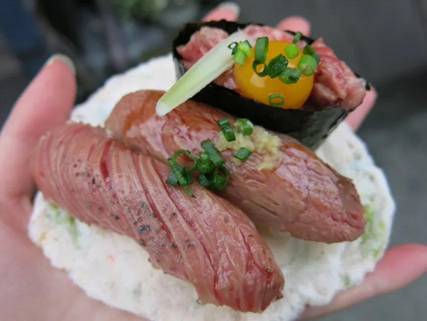 Hida Beef Sushi 手が2つの seared ビーフ寿司を細かく刻んだ青ねぎと小さな葉でトッピングした皿を持っている。その背後には、生のビーフが詰められた海苔に巻かれた寿司があり、その上には生のウズラの卵と青ねぎが飾られている。
