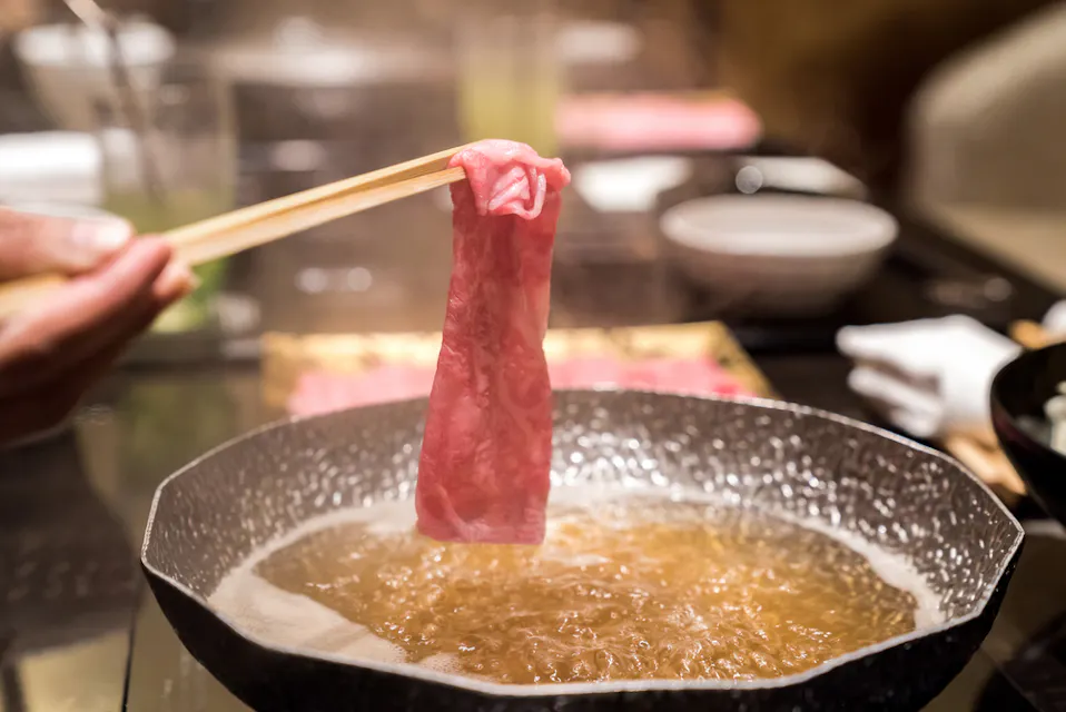Hida Beef Shabu Shabu 鍋の煮汁の上に生肉の薄切りを箸で持つ手。シーンは、レストランの設定でしゃぶしゃぶまたは鍋料理を示しており、背景はぼやけていて他の料理が少し見える。