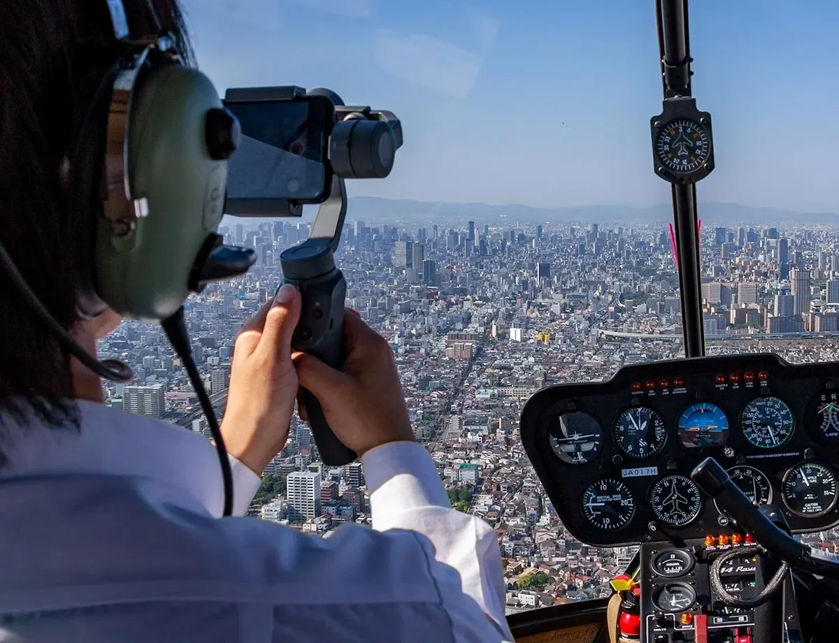 Osaka Helicopter Tour View from inside a helicopter flying over a city; two pilots in headsets are visible, with one filming on a phone. The cockpit dashboard and sprawling urban landscape below are in focus.