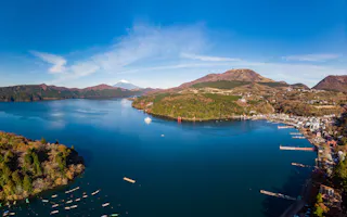 Aerial view of Lake Ashi with deep blue water, surrounded by green hills and mountains; Mount Fuji is visible in the background under a clear blue sky. A small town and boats line the shore.