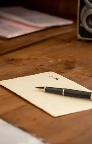 Washi Paper A pen rests on a folded paper atop a wooden desk, with folders, a vintage camera, and a small black cat figurine in the background.