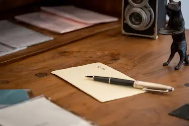 A pen rests on a folded paper atop a wooden desk, with folders, a vintage camera, and a small black cat figurine in the background.