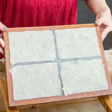 Washi Paper Making A person in a red shirt holds a wooden frame with a mesh screen, displaying four sheets of freshly made, wet paper during a papermaking process.