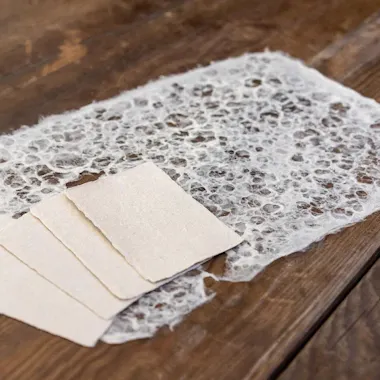 Washi Paper Making A sheet of handmade, textured paper with a lacy, net-like pattern lies on a wooden surface beside three small, rectangular sheets of plain beige paper.