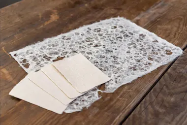 A sheet of handmade, textured paper with a lacy, net-like pattern lies on a wooden surface beside three small, rectangular sheets of plain beige paper.
