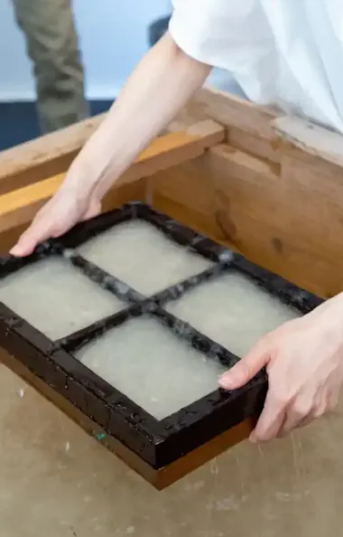 Washi Paper Making A person holds a wooden frame with four sections over a vat of water, demonstrating a traditional papermaking process.