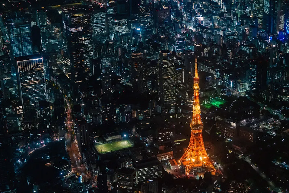Aerial view of a brightly lit urban area at night featuring a prominent illuminated orange tower in the center, surrounded by numerous buildings and streets glowing with city lights.