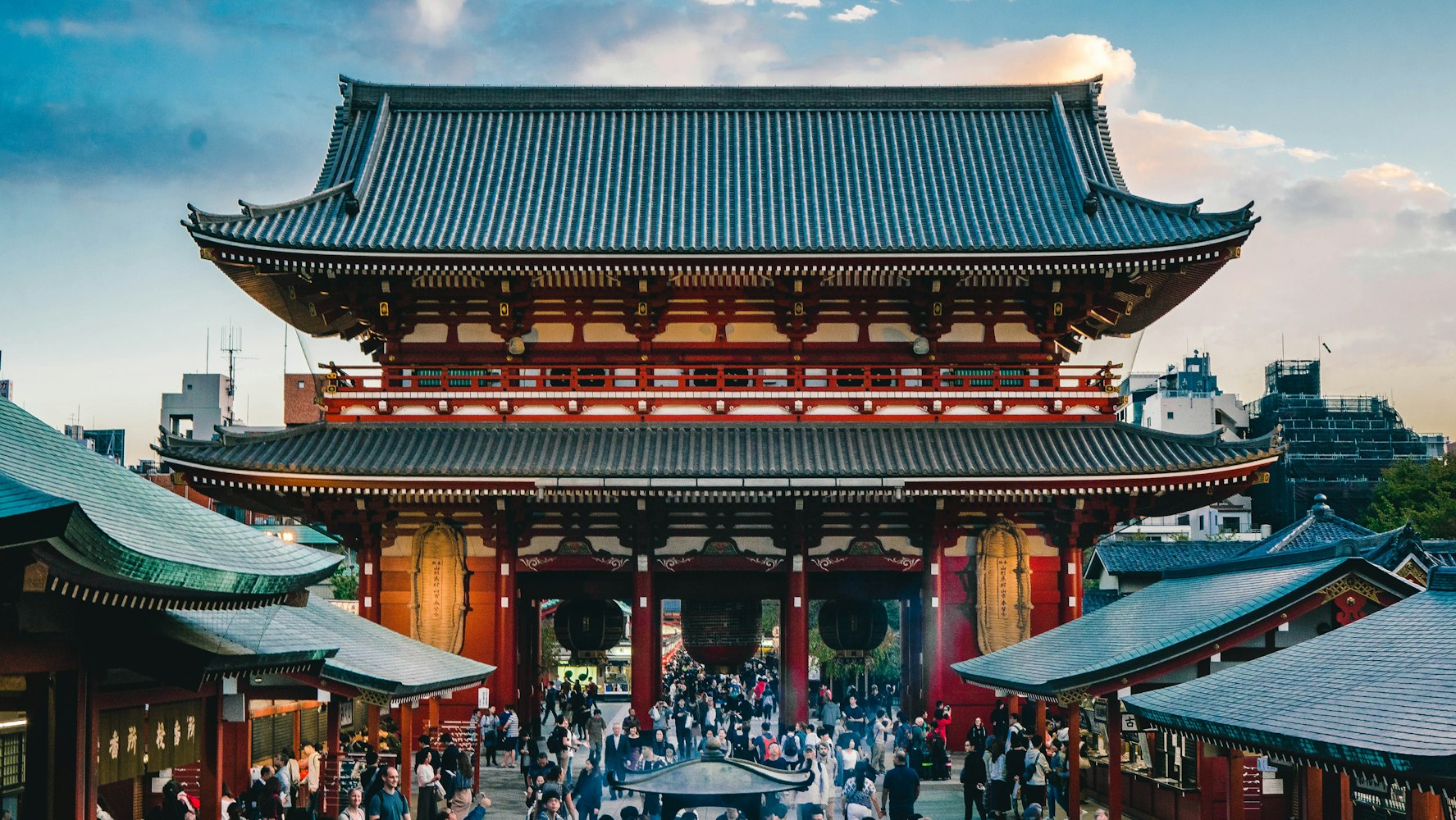 一大群人走過一座傳統日本寺廟的場地，寺廟有一扇大紅白木門，屋頂高高在上。門的兩側有較小的建築，天空部分多雲。