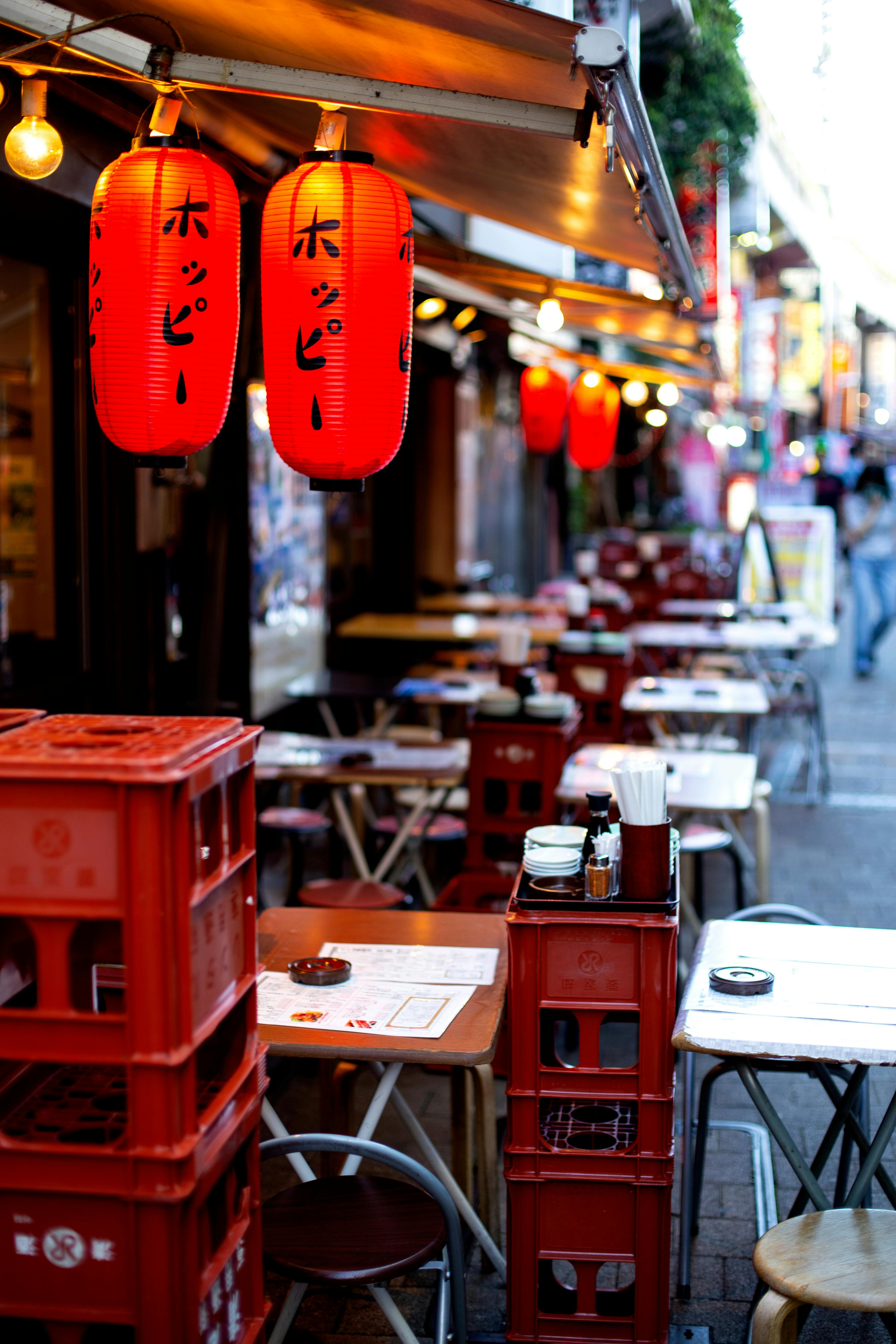 Rows of outdoor tables and chairs line a street, decorated with glowing red Japanese lanterns. Menus and condiments are set on the tables, creating a cozy, inviting atmosphere for dining.