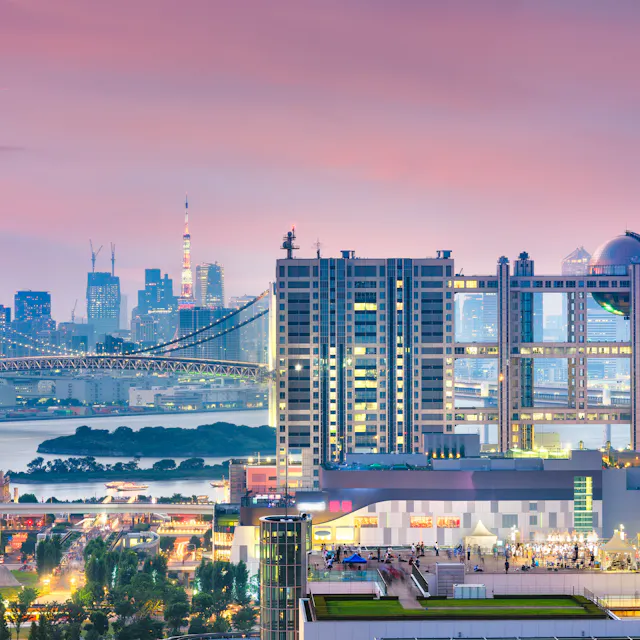 Odaiba Colorful cityscape of Tokyo at sunset, featuring modern high-rise buildings, the Rainbow Bridge, and Tokyo Tower in the background under a pink and purple sky.