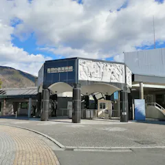 Hakone's Open-Air Museum A modern museum entrance under a partly cloudy sky, featuring curved, dark pillars supporting a detailed white archway adorned with classical relief sculptures. The surrounding area includes a cobblestone walkway, trees, and a backdrop of distant mountains.