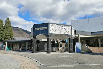 Hakone's Open-Air Museum A modern museum entrance under a partly cloudy sky, featuring curved, dark pillars supporting a detailed white archway adorned with classical relief sculptures. The surrounding area includes a cobblestone walkway, trees, and a backdrop of distant mountains.