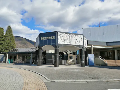 Hakone's Open-Air Museum A modern museum entrance under a partly cloudy sky, featuring curved, dark pillars supporting a detailed white archway adorned with classical relief sculptures. The surrounding area includes a cobblestone walkway, trees, and a backdrop of distant mountains.