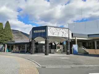 A modern museum entrance under a partly cloudy sky, featuring curved, dark pillars supporting a detailed white archway adorned with classical relief sculptures. The surrounding area includes a cobblestone walkway, trees, and a backdrop of distant mountains.