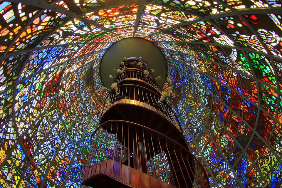 円形の塔の内部にある、くねくねとした木製の螺旋階段が上に昇ります。複雑で多色のステンドグラスで飾られています。鮮やかな色合いが、光がガラスを通過する際に万華鏡のような効果を生み出し、構造内部にカラフルなパターンを投影します。