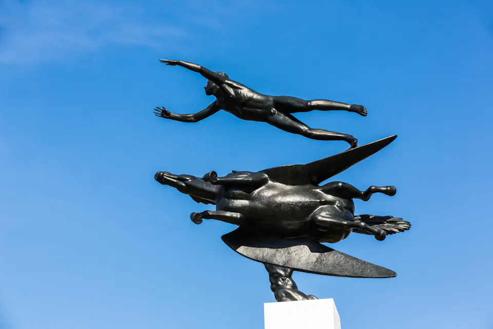 彫刻は、人間の形が翼のある馬の上を飛んでいる動的なシーンを描いています。両方の形は、まるで動いているかのように水平方向に向いており、透き通った青空を背景にしています。このアート作品は、白い台座の上に設置されています。