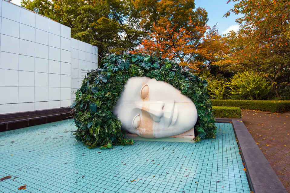 穏やかな顔の大きな白い彫刻が、浅いタイルのプールの横に横たわっています。彫刻の髪は緑の葉で構成され、周囲の木々や低木と溶け込んでいます。この地域は静かで、背景には秋の色合いがあります。